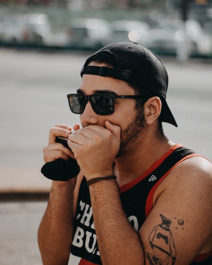 Young man with sunglasses playing harmonica outdoors on a sunny day. Casual and expressive.