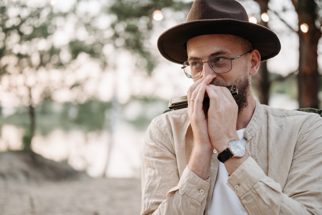 Portrait of a bearded man wearing a fedora, playing harmonica outside with blurred background.