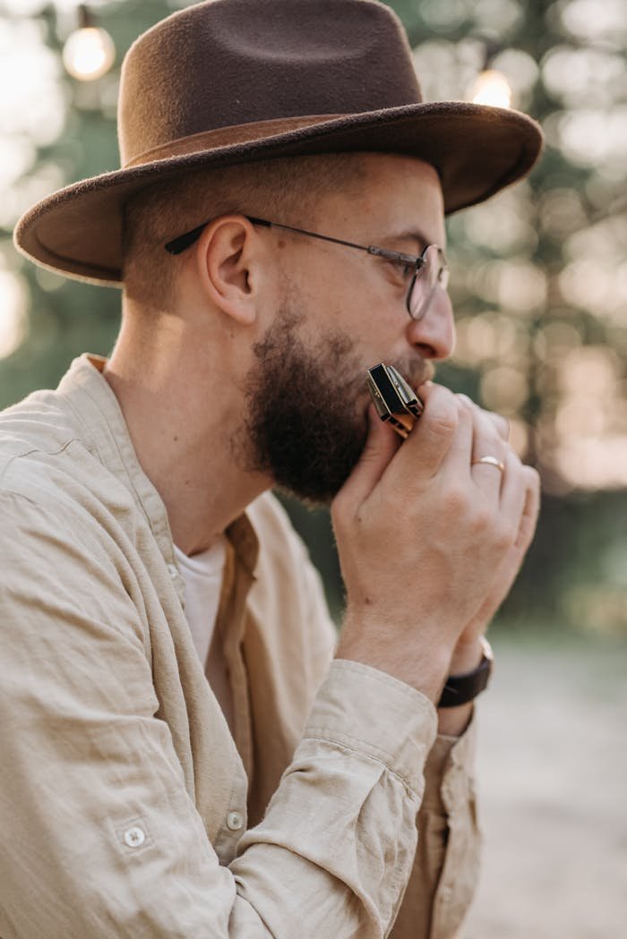 Side view of a man wearing a hat playing harmonica outdoors, captured during the day.